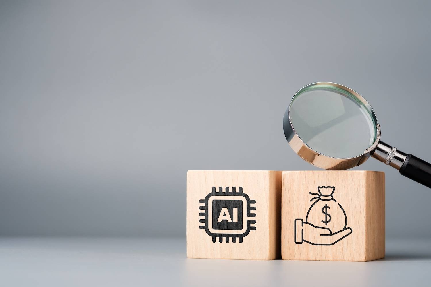 Magnifying glass hovers over wooden blocks with AI and money bag symbols on a gray background.
