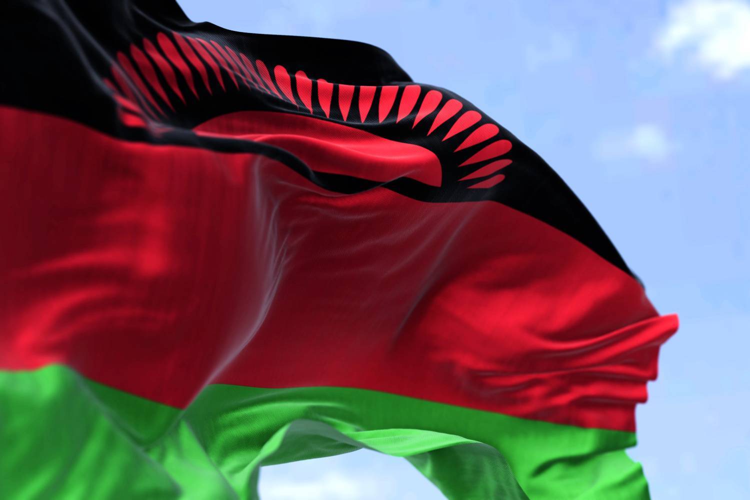 Malawi flag waving with black, red and green stripes under a blue sky.