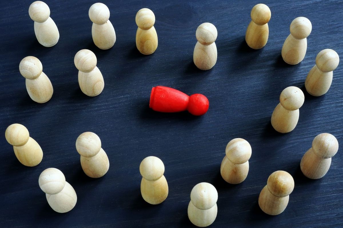 Many small beige wooden figures stand surrounding one red figure laying on its side.