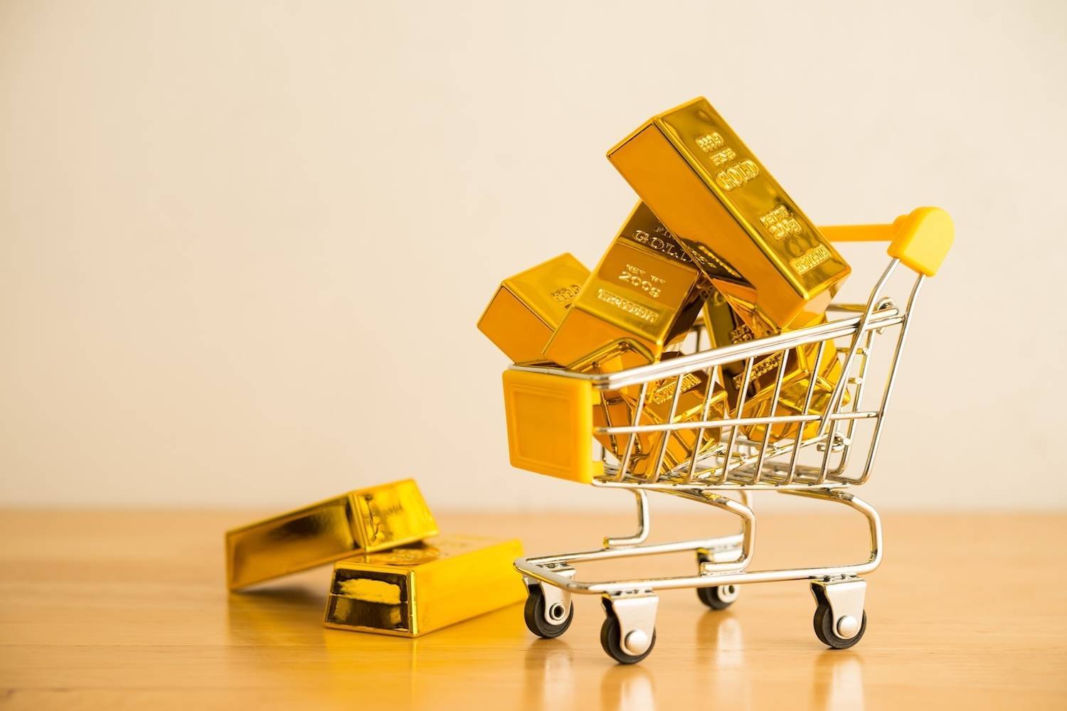 Mini shopping cart overflowing with gold bars on a wooden surface.