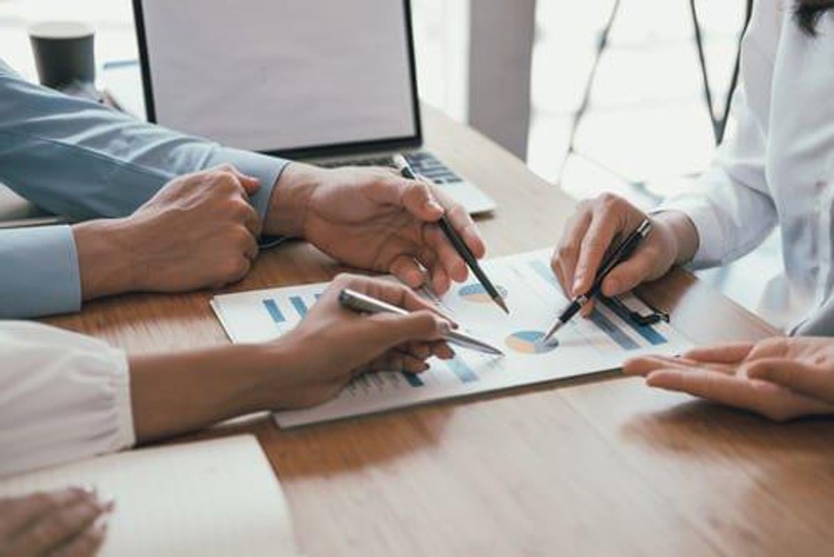 multiple people sitting around a desk each point a pen at a pie chart on a paper