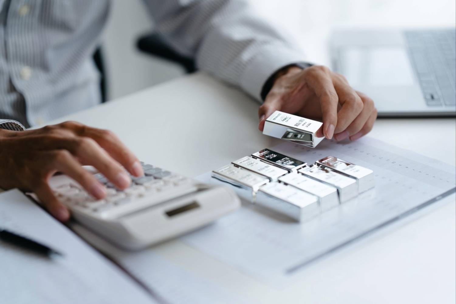 Person calculating with a calculator and examining silver bullion on a desk.