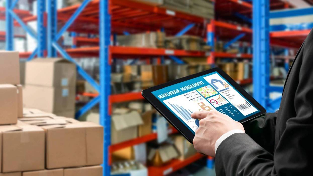 Person holding a tablet with "warehouse management" screen in front of large shelves and stacks of boxes.