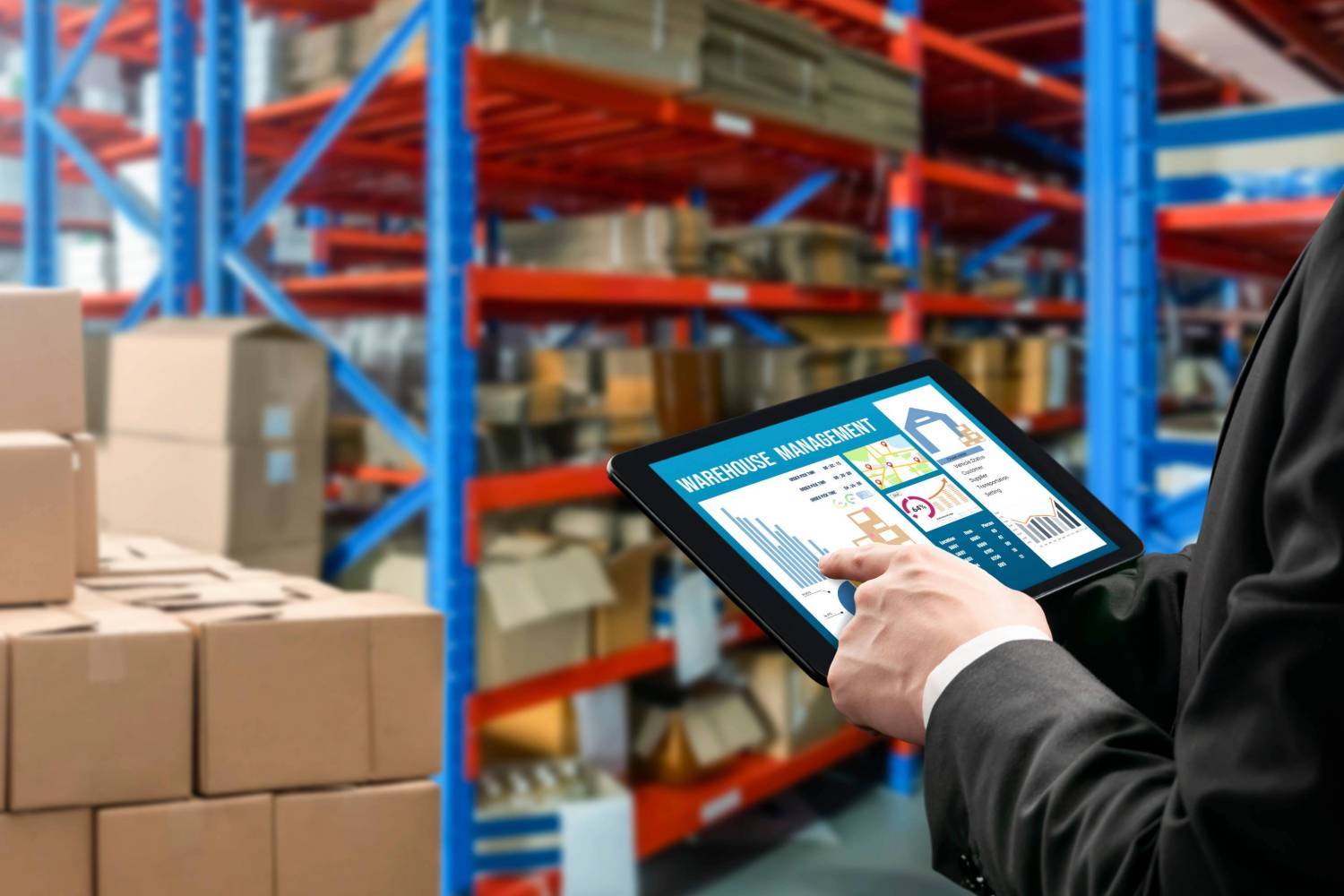 Person holding a tablet with "warehouse management" screen in front of large shelves and stacks of boxes.