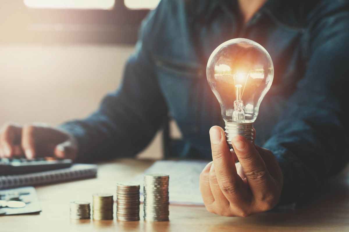 person holding lit light bulb over desk next to ascending stacks of coins