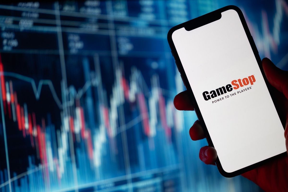 Person holding phone that says "GameStop" in front of stock trading charts.