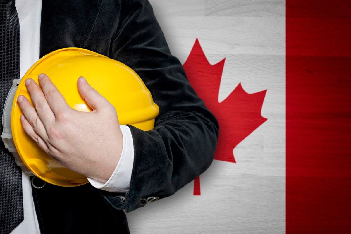 Person in suit holding yellow engineer helmet stands in front of Canada flag.