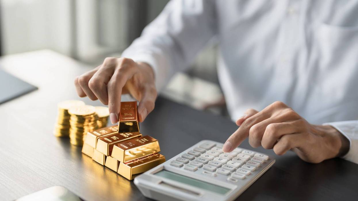 Person in white shirt using a calculator beside stacked gold bars and coins on a dark table.