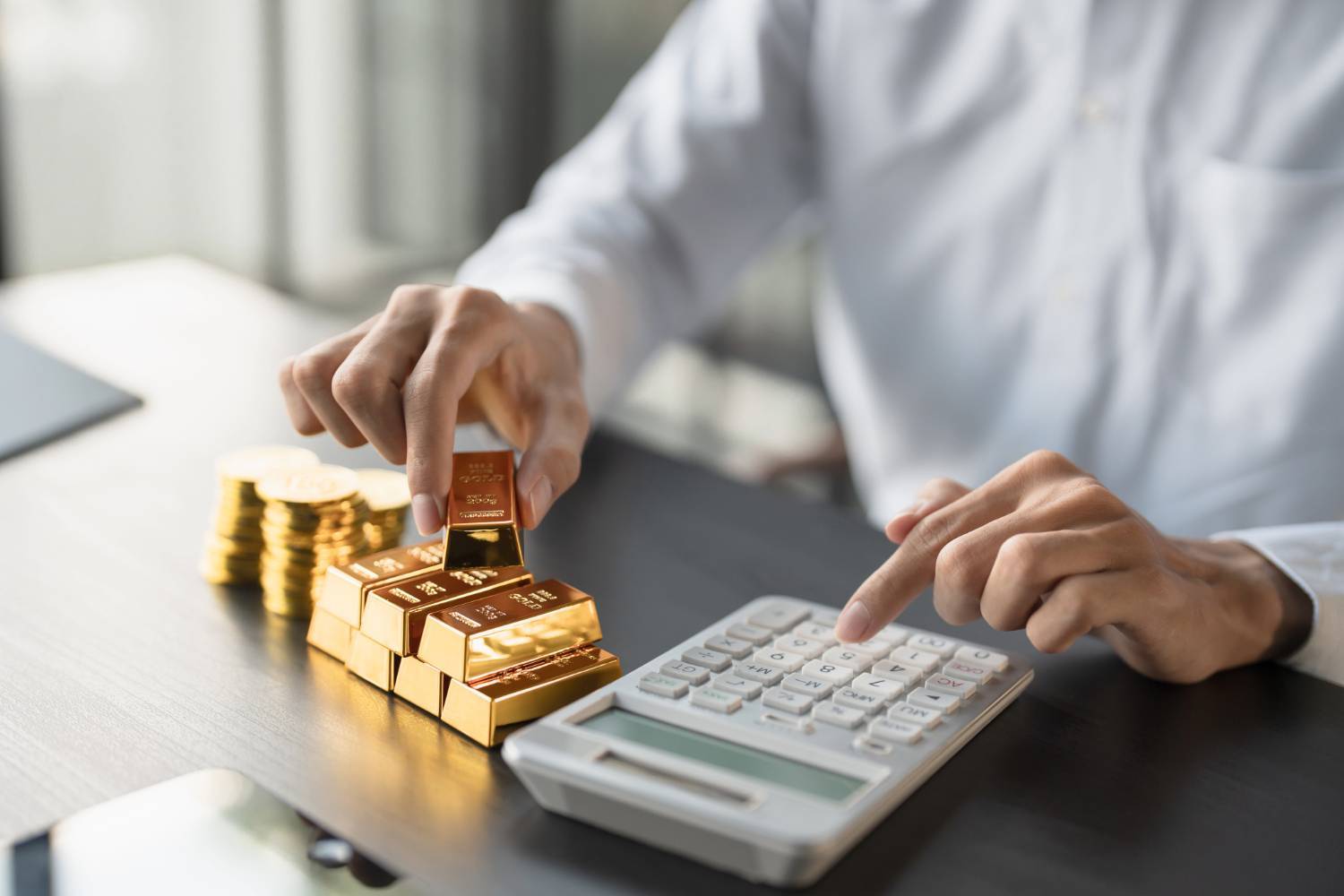 Person in white shirt using a calculator beside stacked gold bars and coins on a dark table.