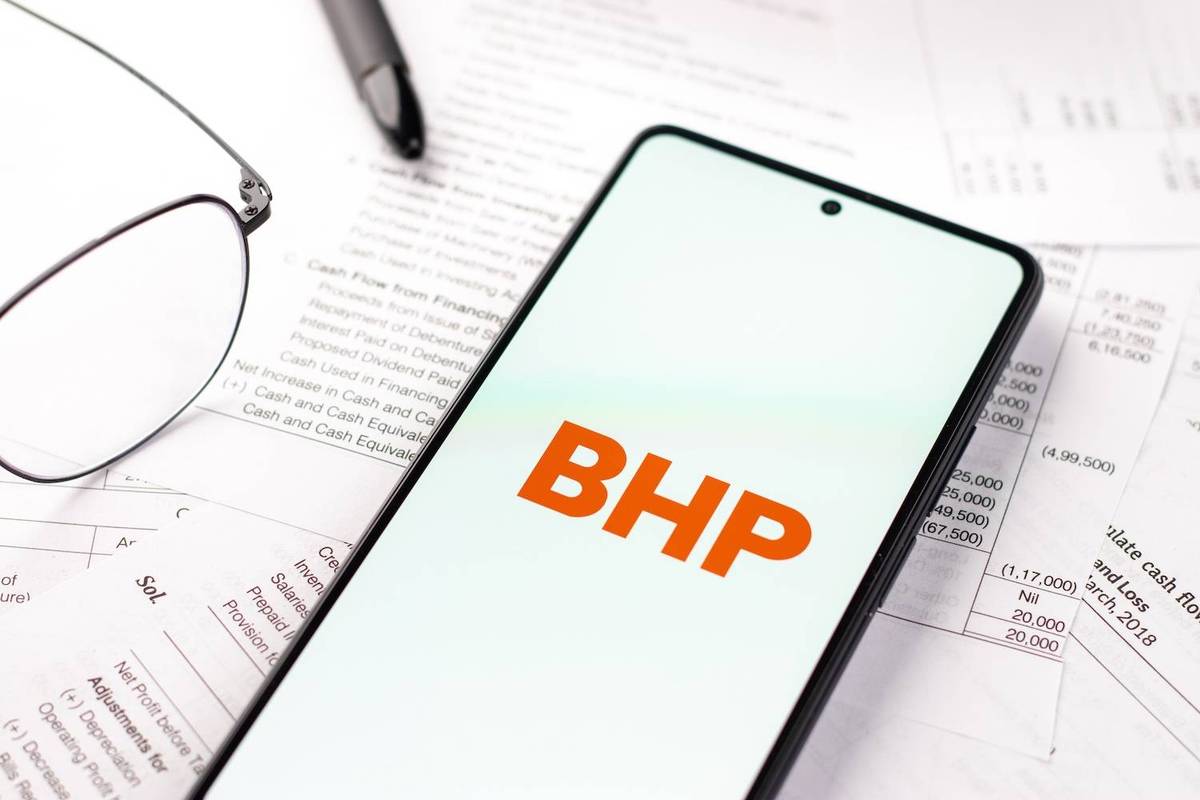 Phone displaying "BHP" on financial documents with glasses and pen.