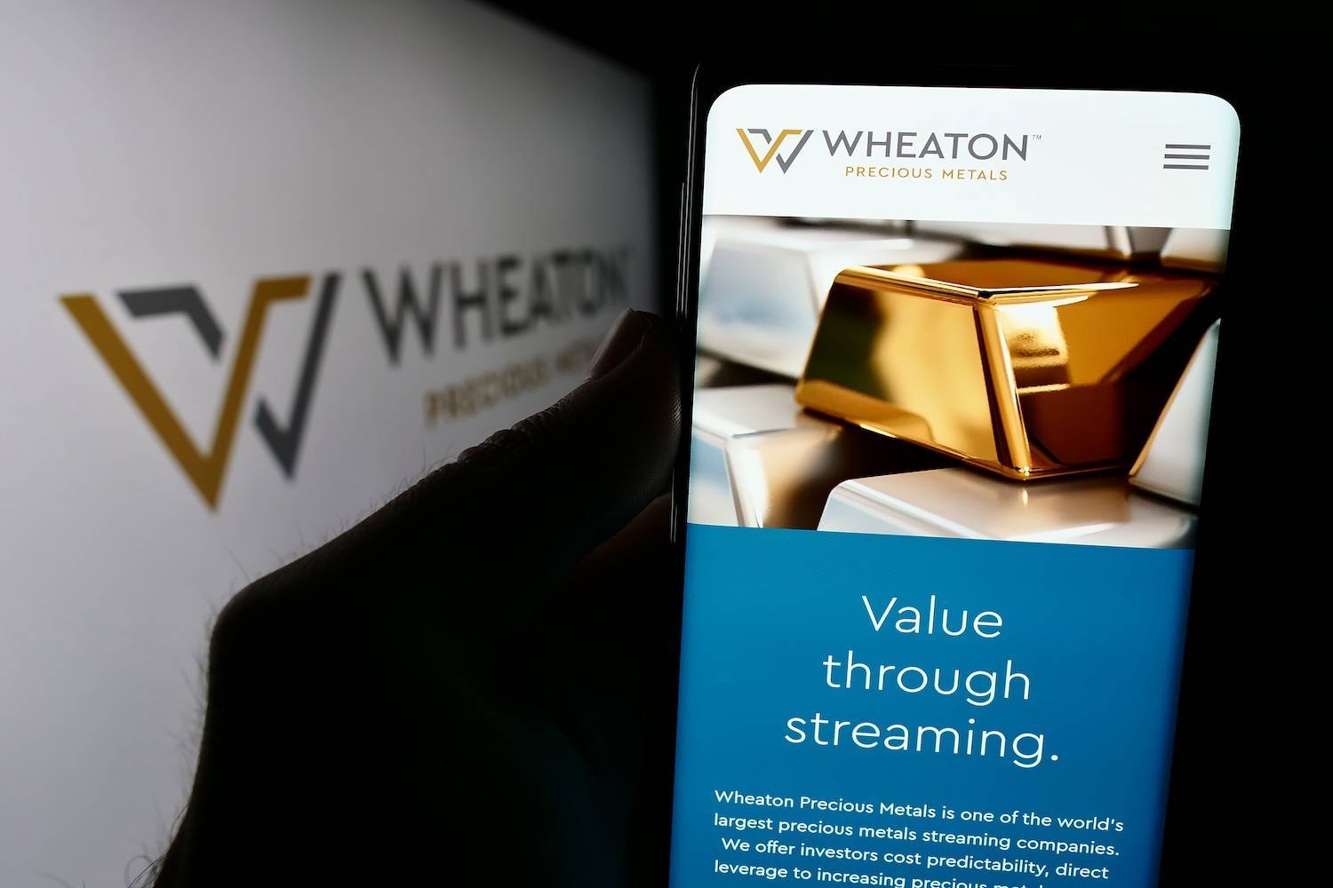 Phone displaying Wheaton Precious Metals website with gold bars on screen; logo and text in background.
