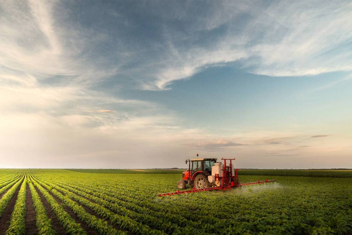 Red tractor sprays lush green field under a blue sky with wispy clouds.