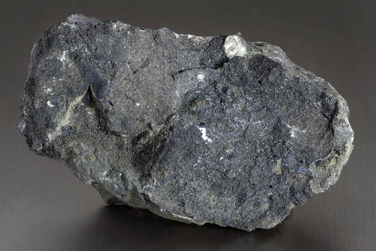 Rough, dark gray sedimentary phosphate rock with jagged texture sits on a smooth, dark gray surface.