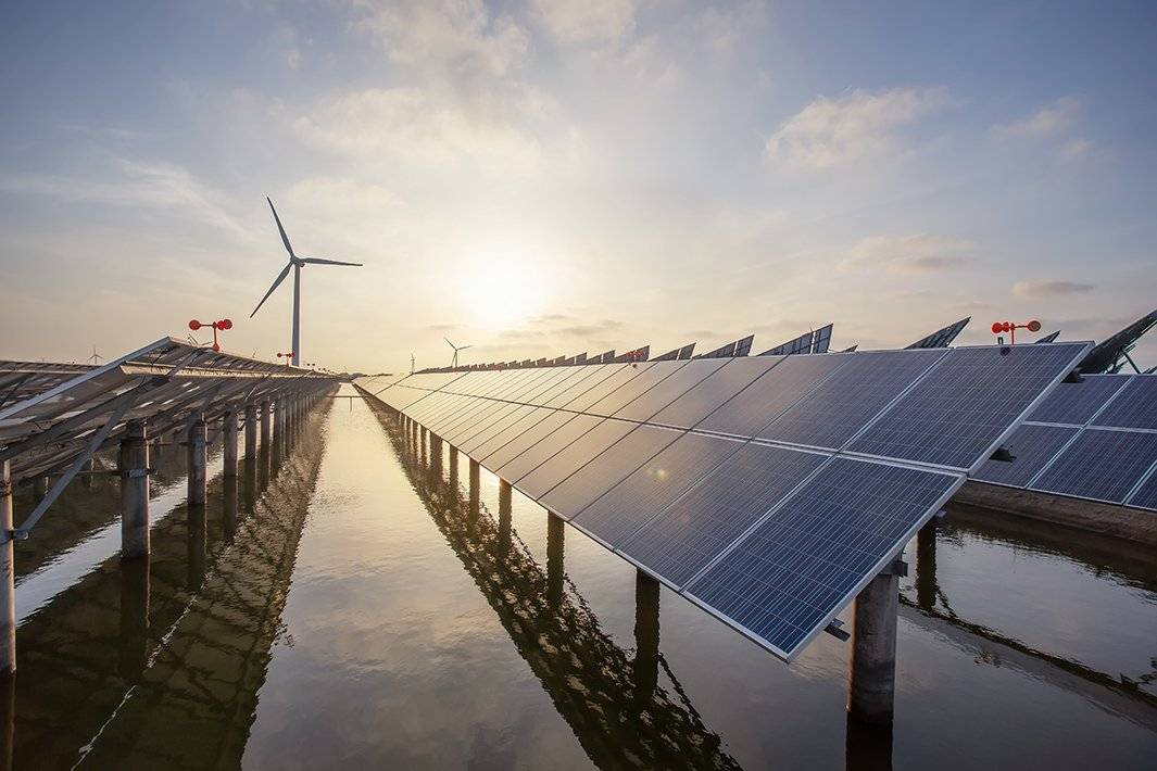 Rows of solar panels and wind turbines over water with a setting sun in the background.