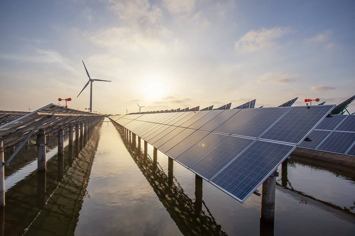 Rows of solar panels and wind turbines over water with a setting sun in the background.