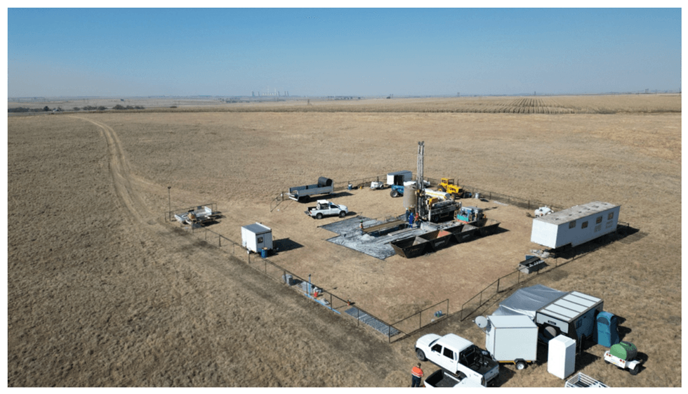 Rural drilling site with equipment, trucks, and trailers in a fenced area, surrounded by fields.