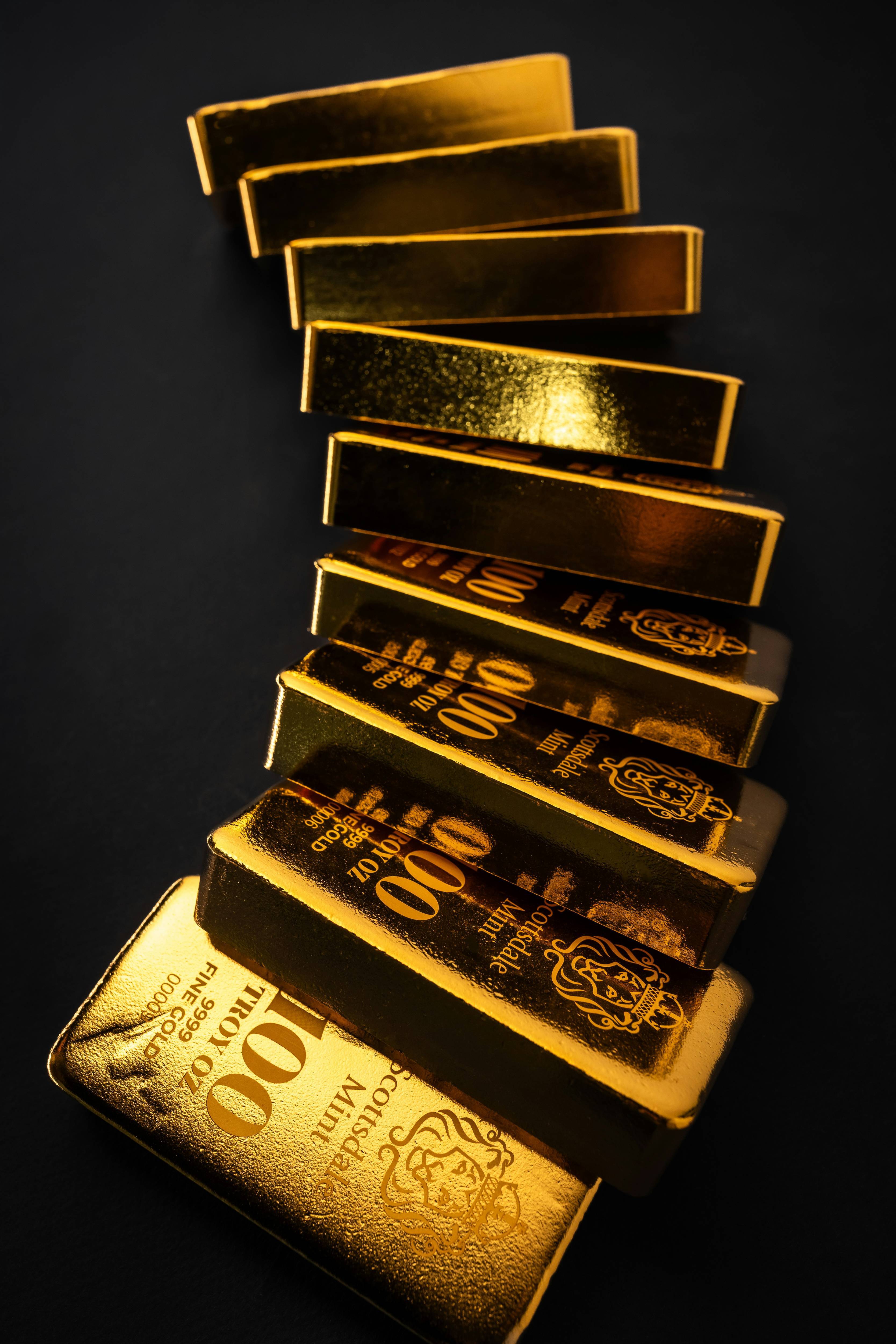 Seven gold bars stacked diagonally over a dark background, text reads: 100 TROY OZ, 999.9 FINE GOLD.