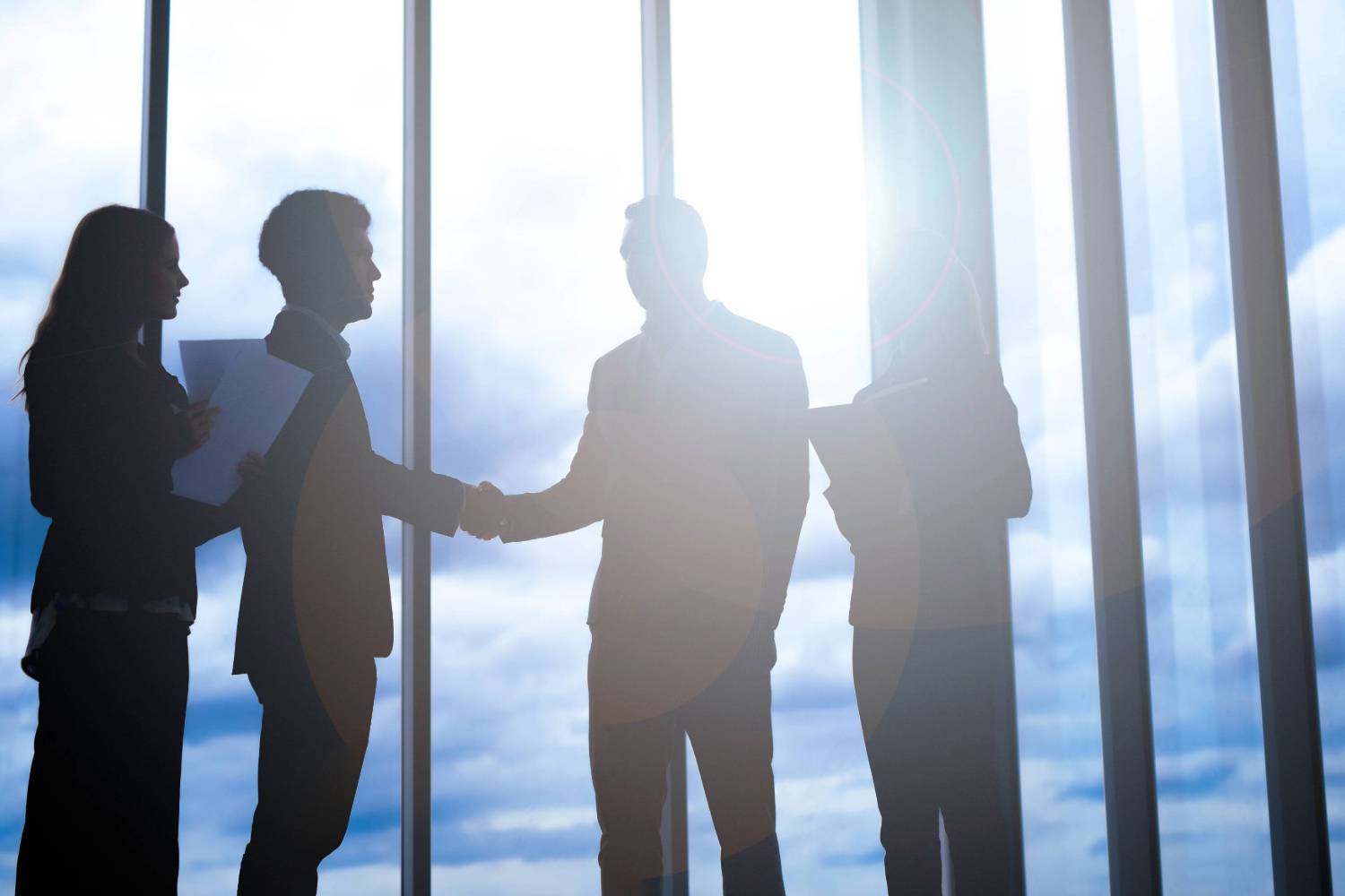 Silhouettes of four people, two shaking hands, stand in front of large windows with a bright sky background.