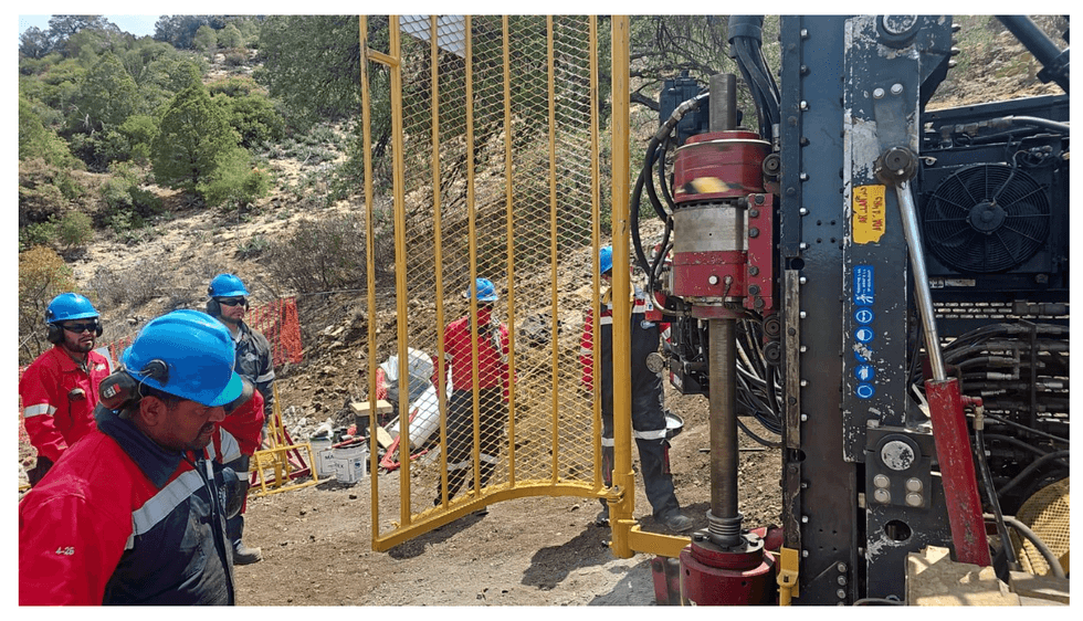 Silver Dollar Resources workers in blue helmets and red uniforms operate machinery with a yellow gate on a rocky hillside.