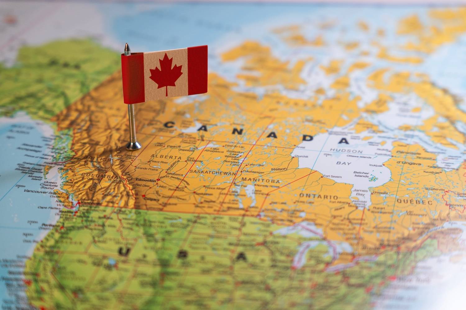 Small Canadian flag pin stands over a colorful map of Canada with visible geographical names.