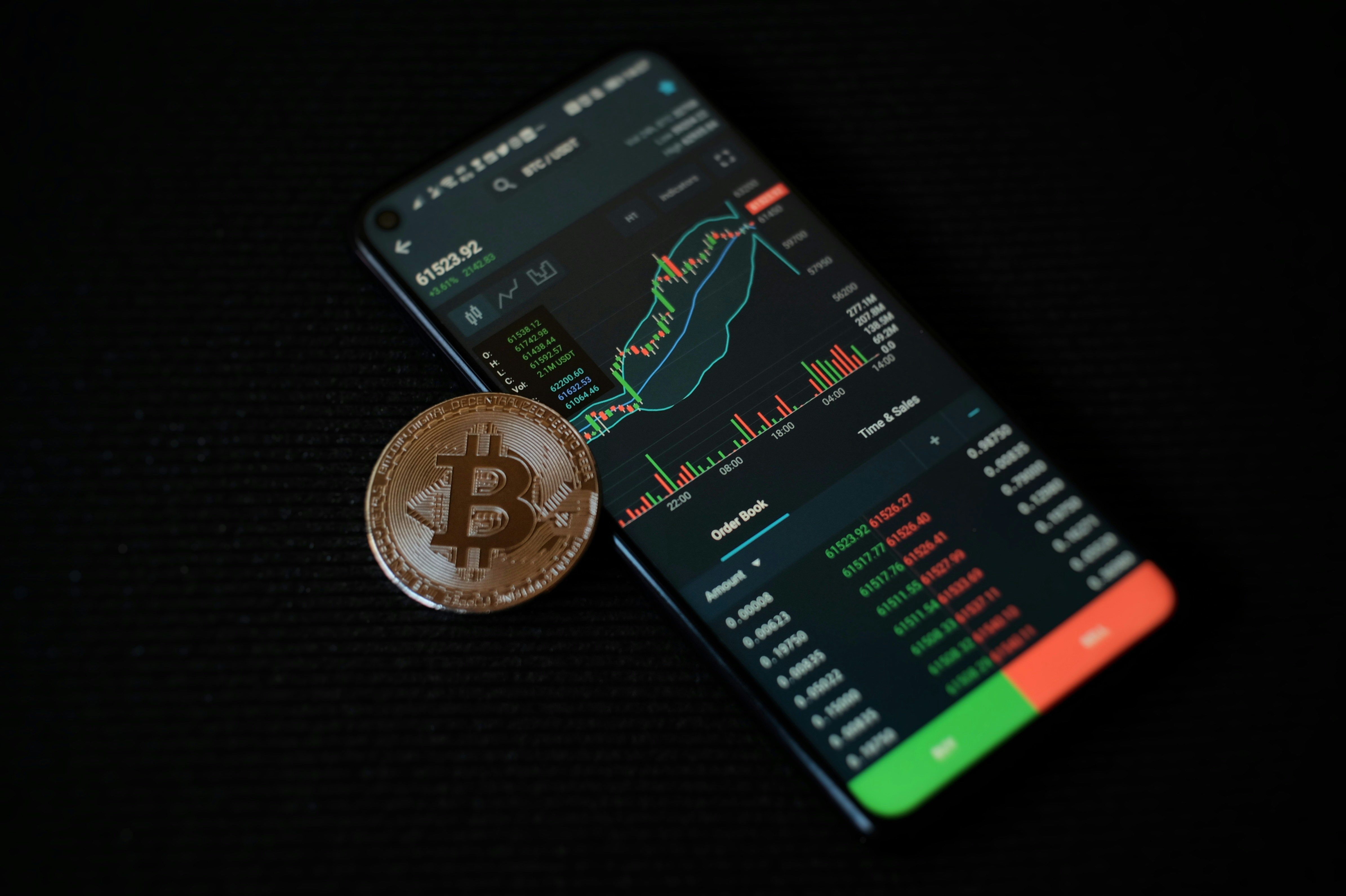 Smartphone displaying crypto charts with a Bitcoin coin on a black background.