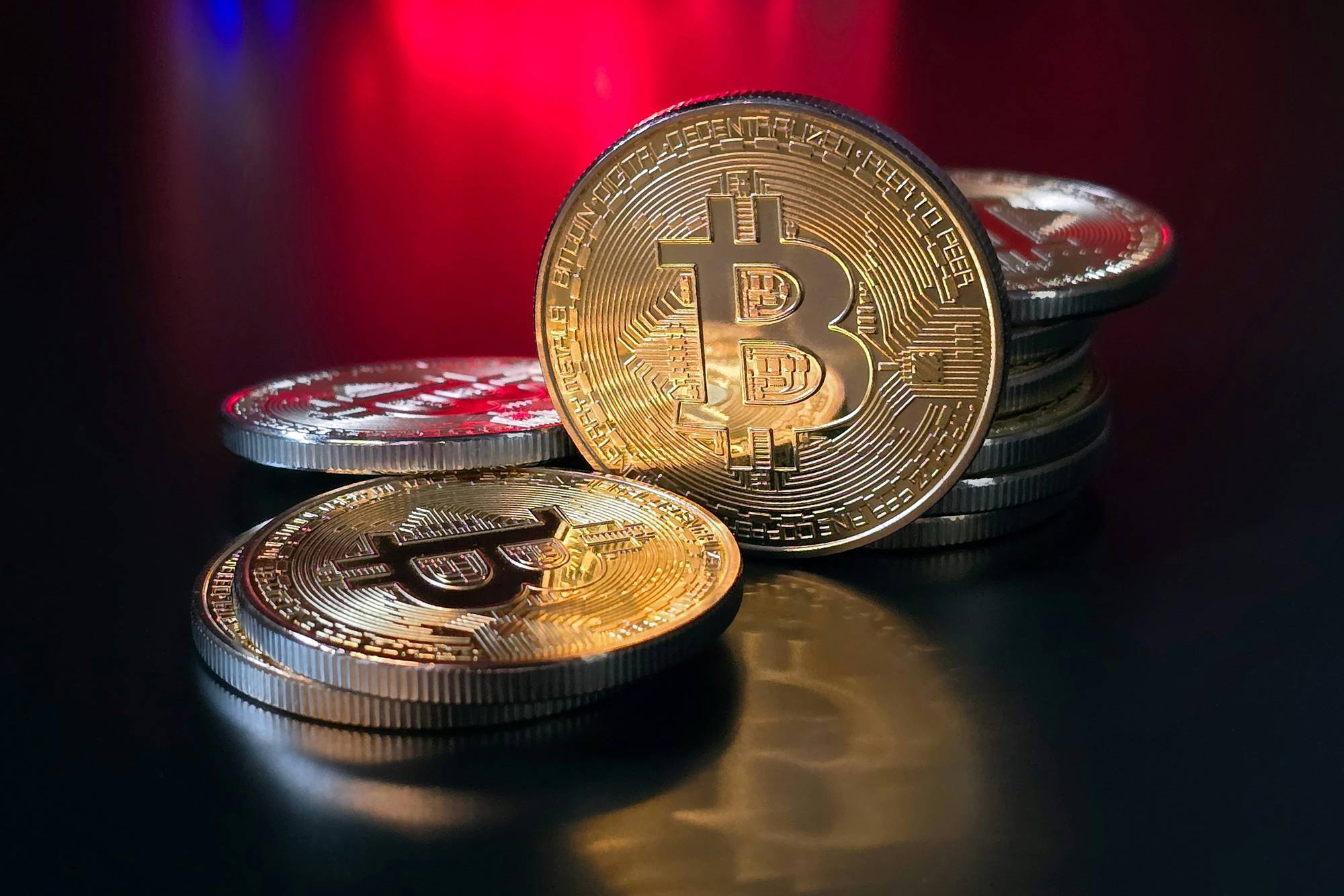 Stack of gold and silver Bitcoin coins on reflective black surface with red and blue background.