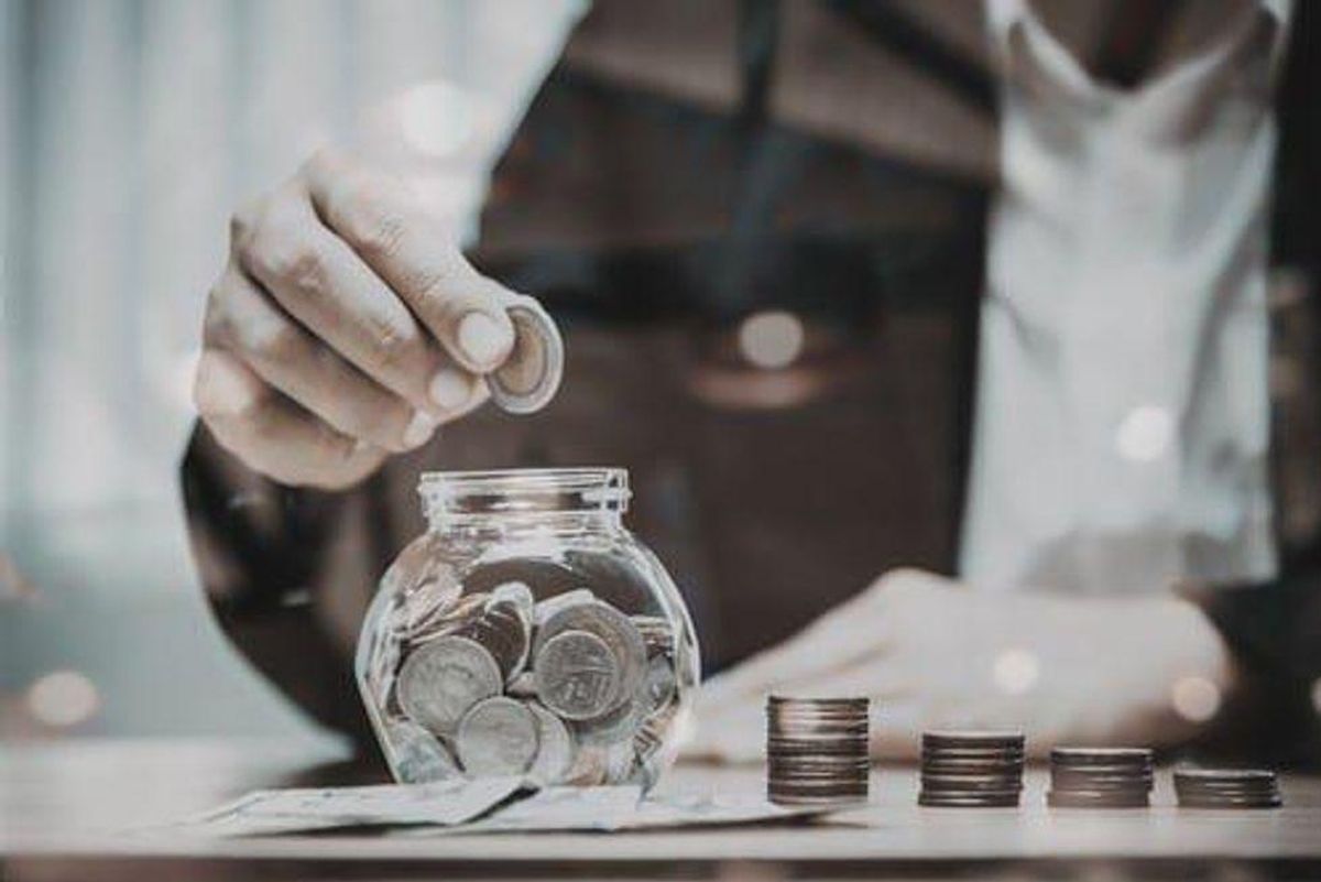stacks of coins are on a table near a jar nearly filled with coins. a person is about to place another coin in the jar.