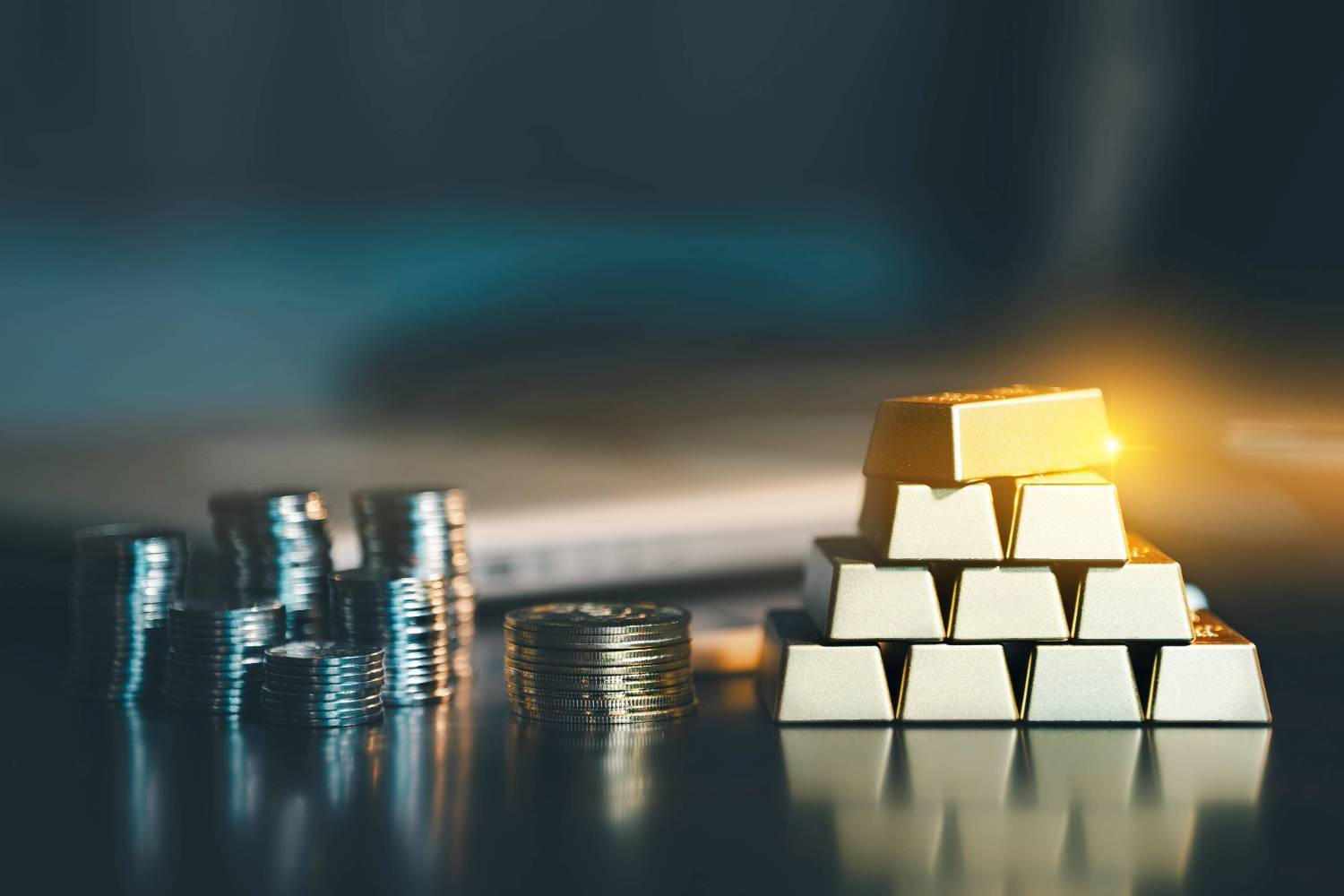 Stacks of silver coins and a pyramid of gold bars on a dark reflective surface.
