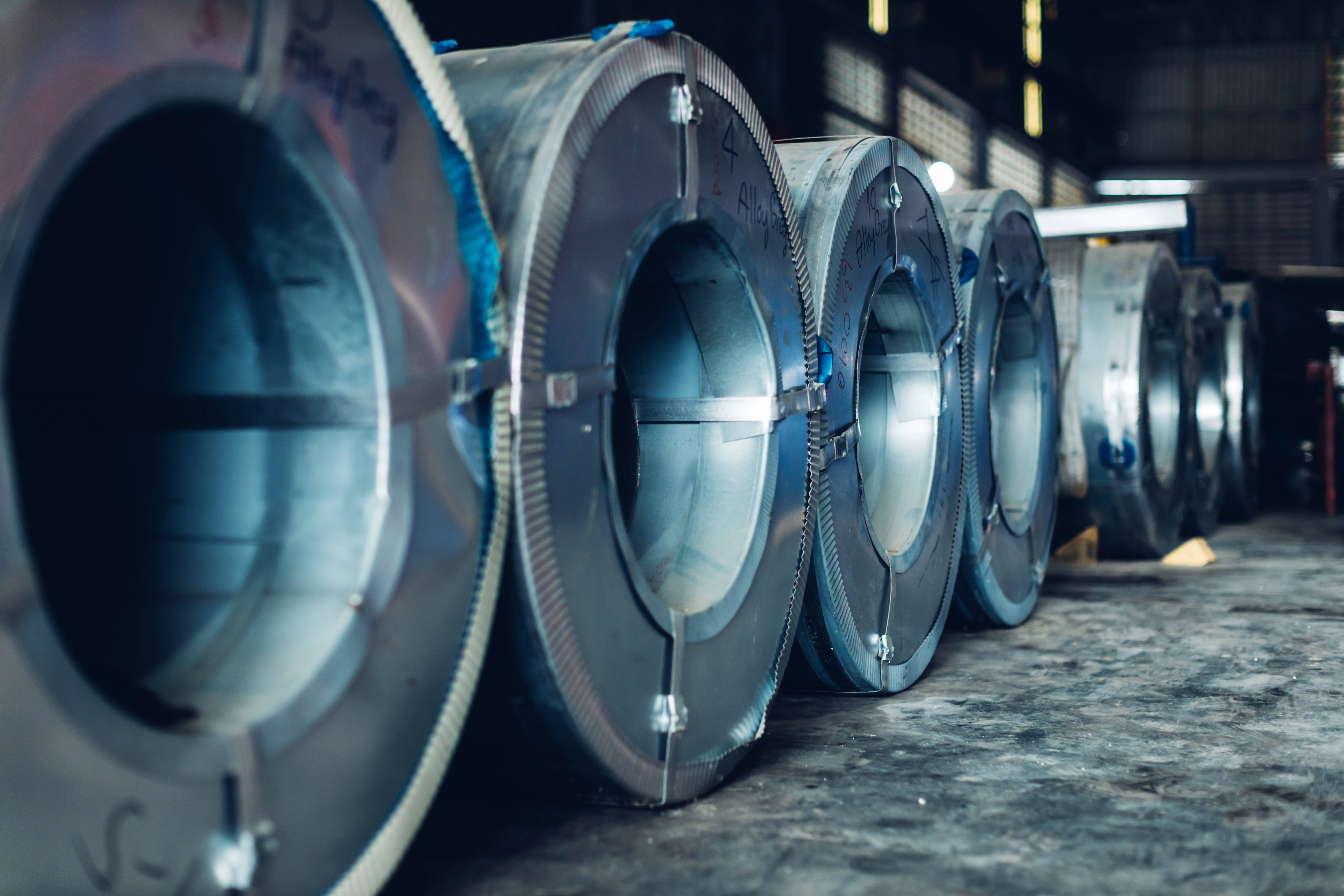 Steel rolls lined up on warehouse floor, with faint text and lighting creating reflections on the surfaces.