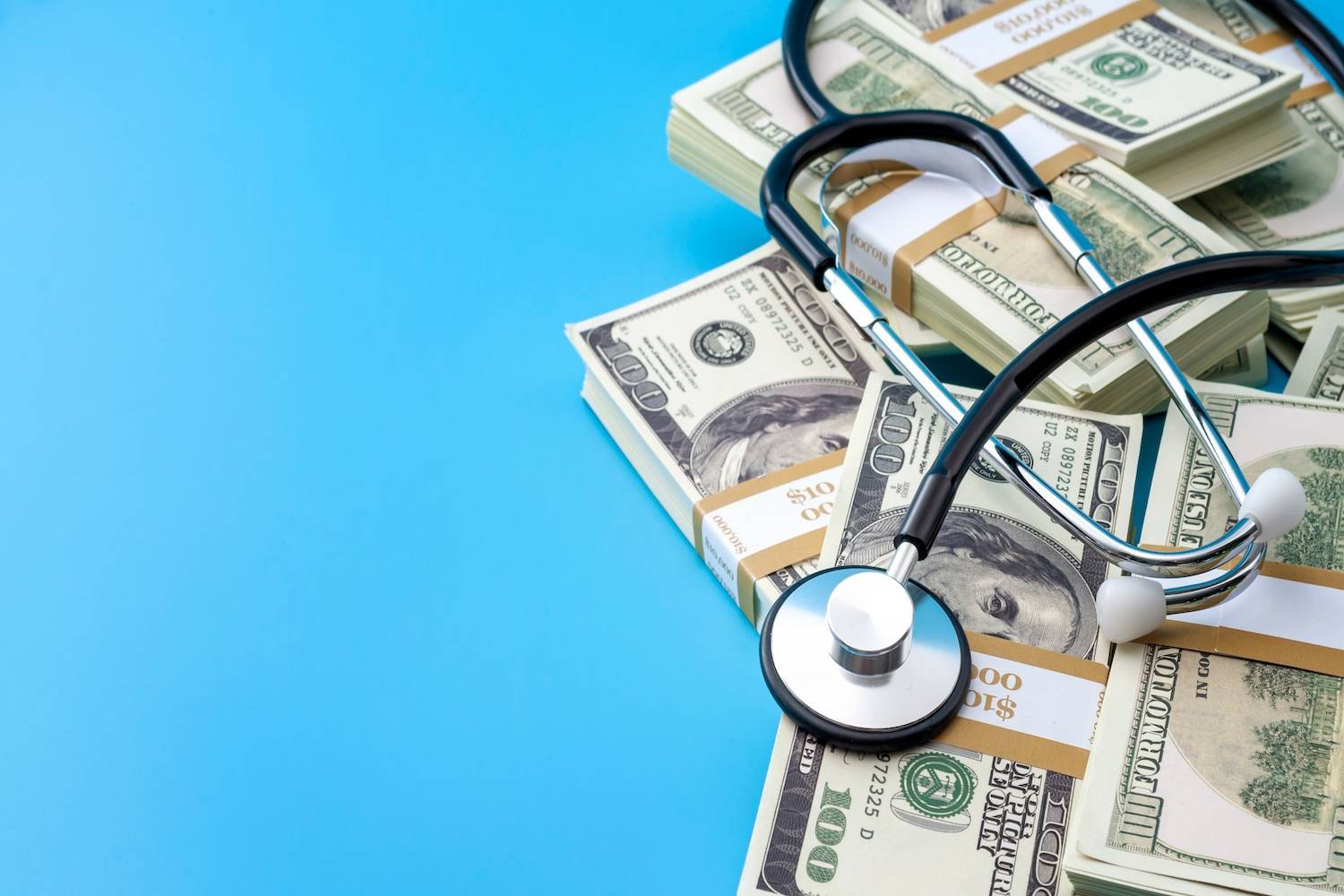 Stethoscope rests on stacks of US money over a bright blue background.