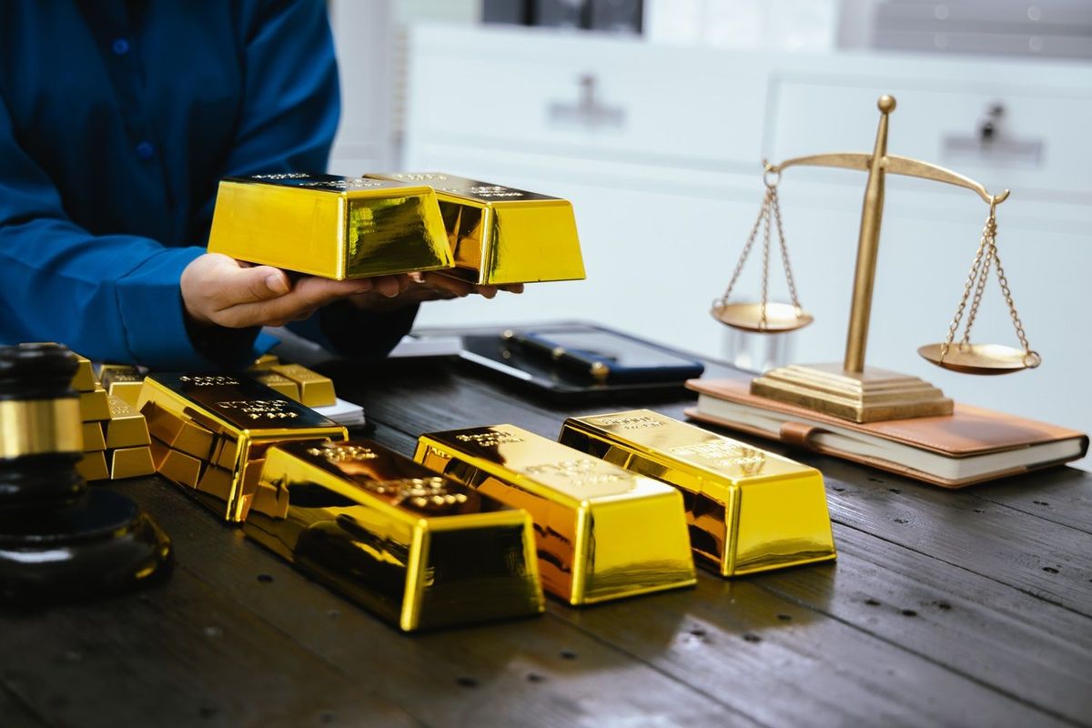 Table with gold bullion bars in varying sizes, a scale and a judge's gavel. A person holds one bar in each hand above the table.