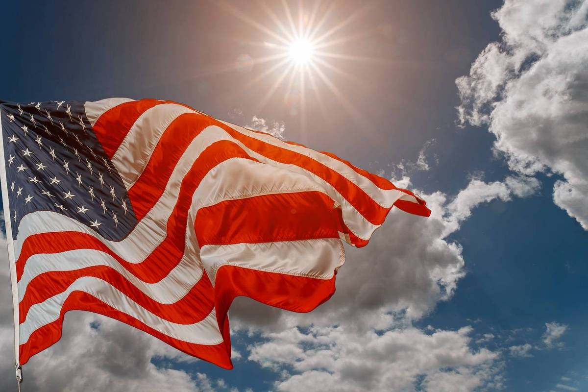 The American flag waves under a bright sun with scattered clouds in a blue sky.