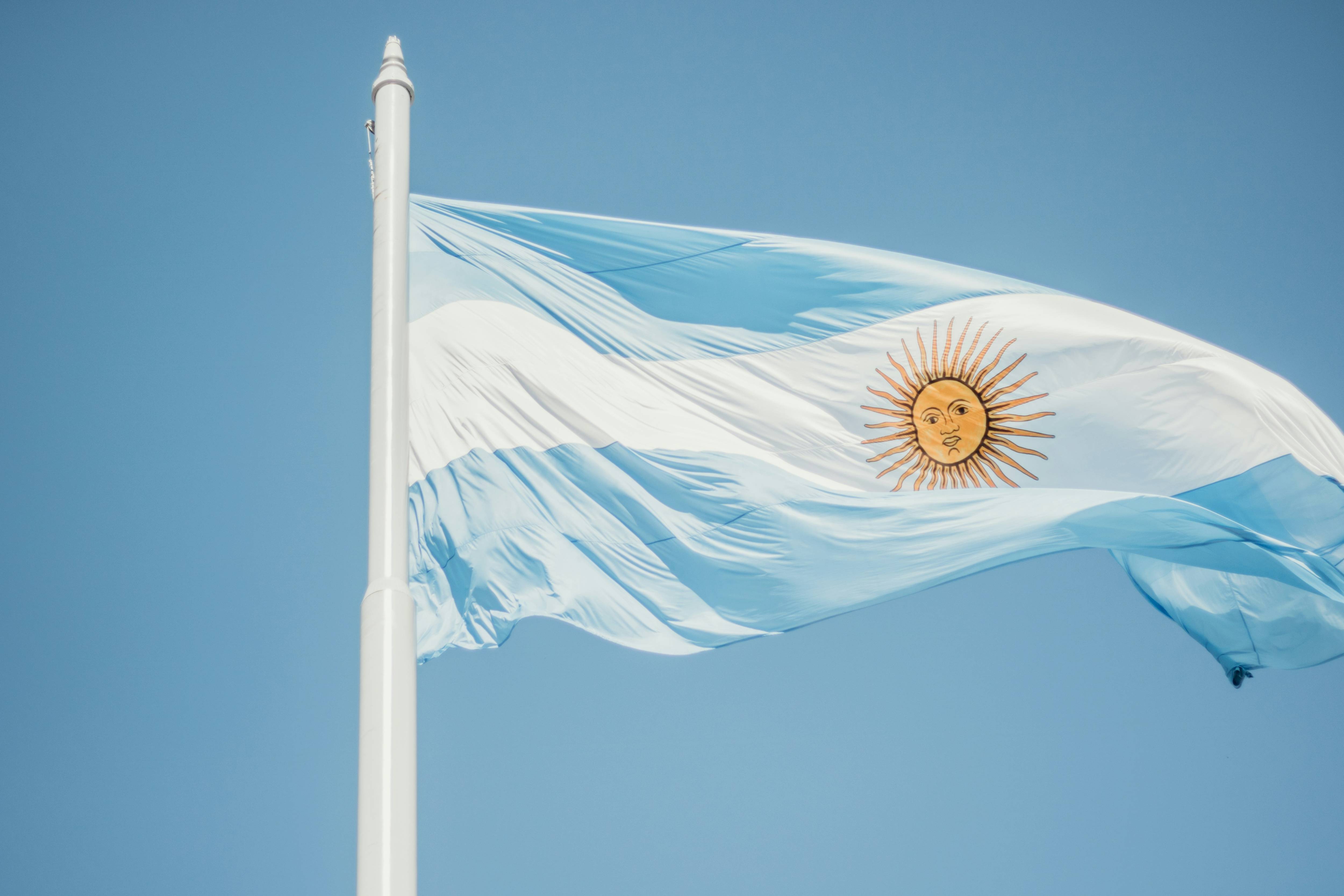 The Argentine flag with a sun emblem waves on a white pole against a clear blue sky.