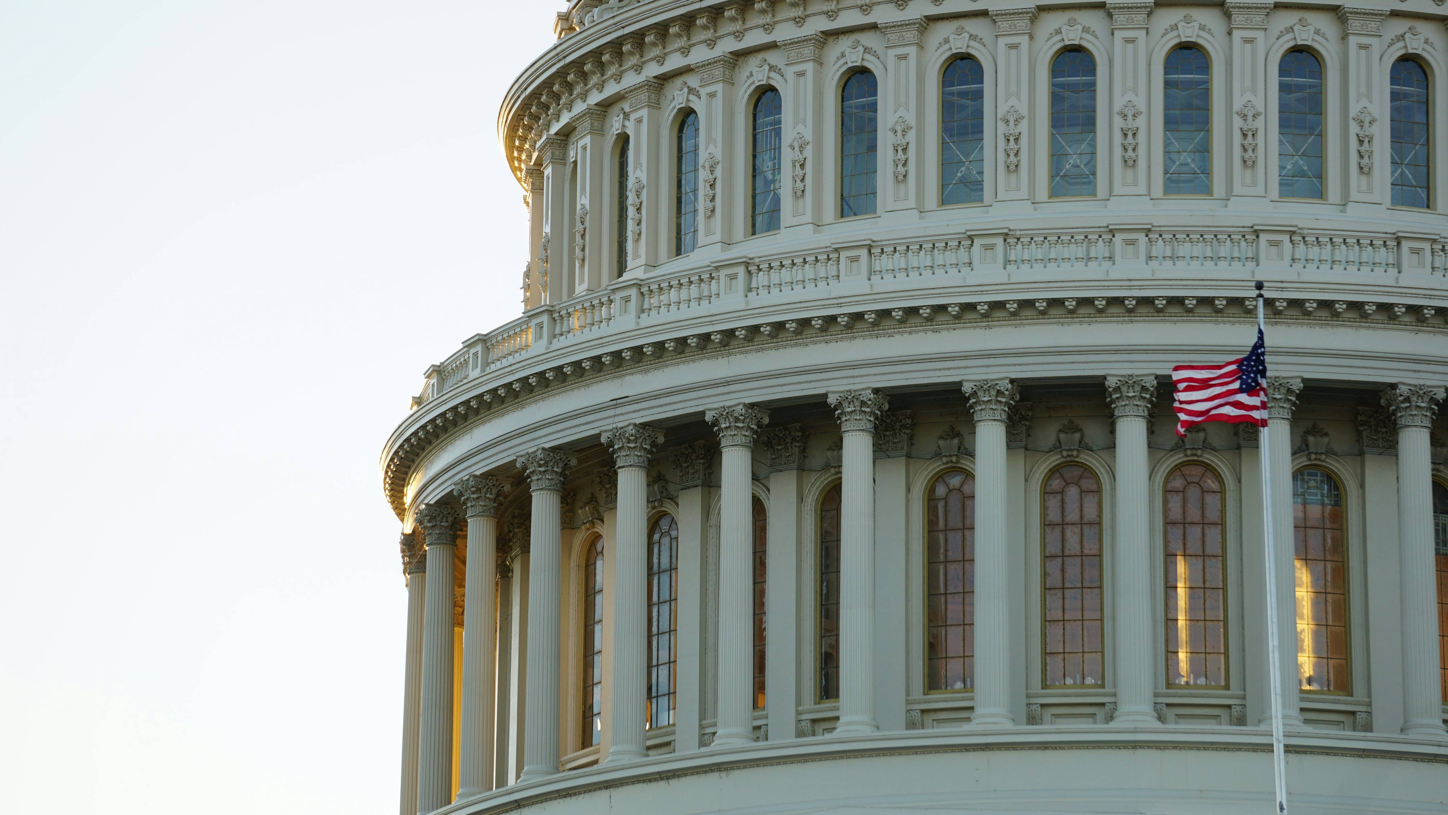 The image shows the U.S. Capitol dome with a waving American flag in front of tall, arched windows.