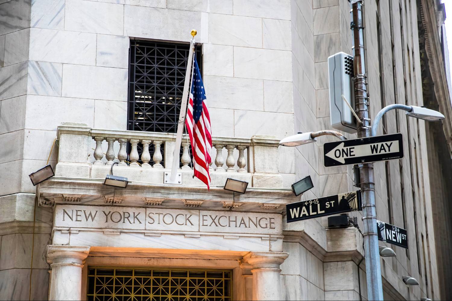 The New York Stock Exchange entrance with a US flag, street signs for Wall Street and a "one way" sign.