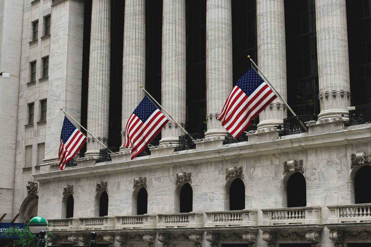Three American flags hang over a stone building with tall columns and arched windows.
