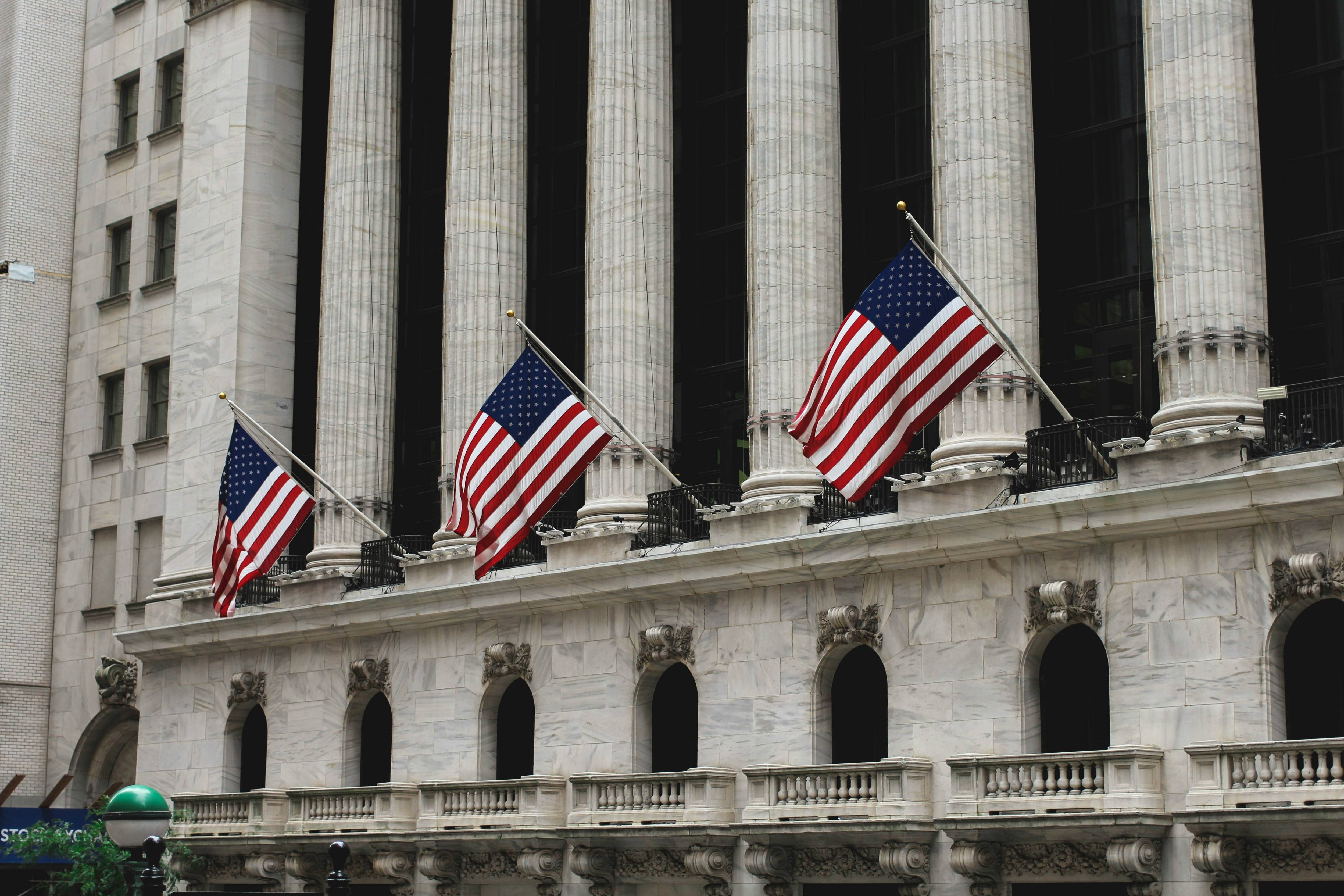 Three American flags hang over a stone building with tall columns and arched windows.