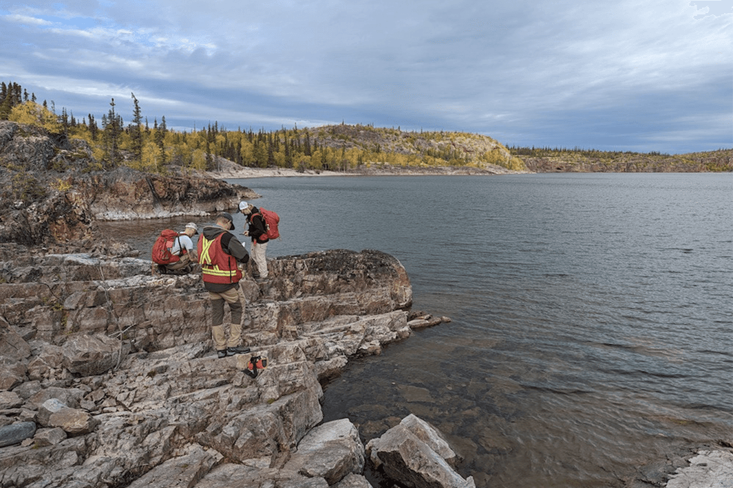 Three geologists in safety vests stand on rocky terrain beside a lake under a cloudy sky with trees in background.