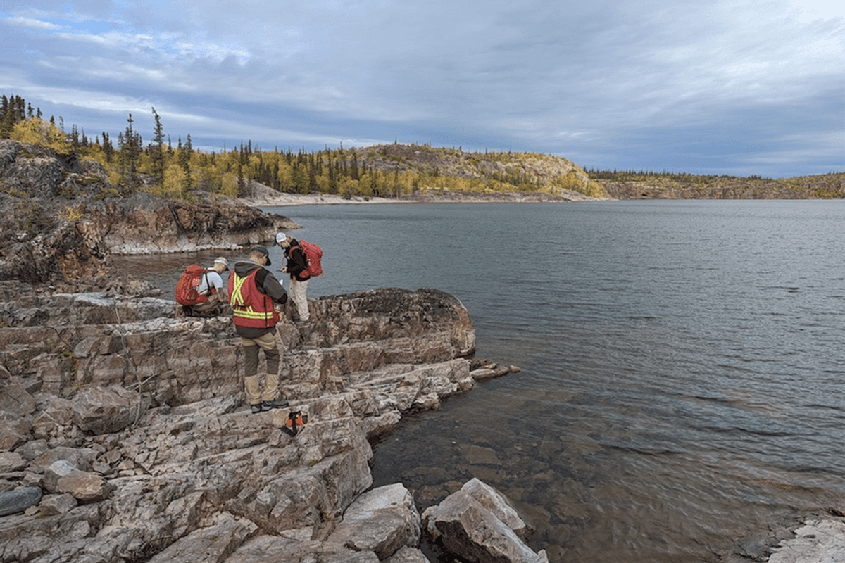 Three geologists in safety vests stand on rocky terrain beside a lake under a cloudy sky with trees in background.