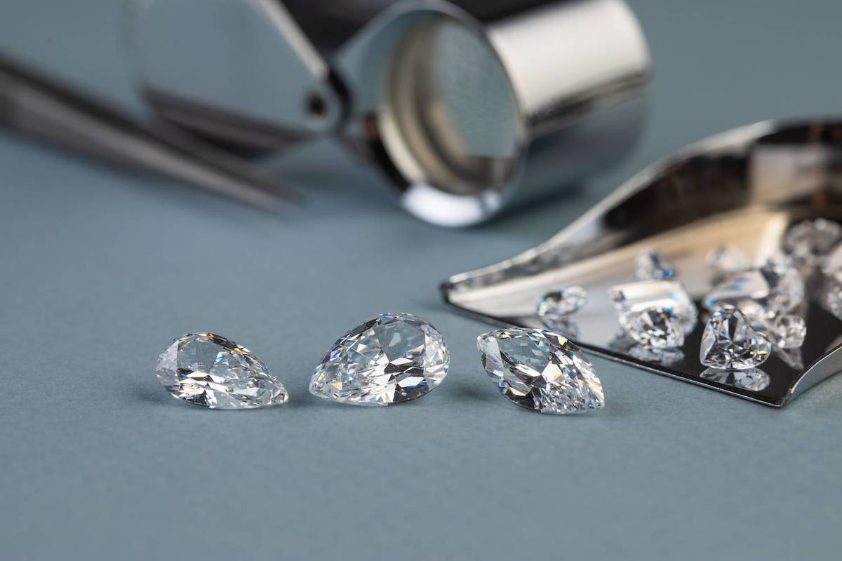 Three pear-cut diamonds on a blue surface with a magnifying loupe and tray of more diamonds nearby.