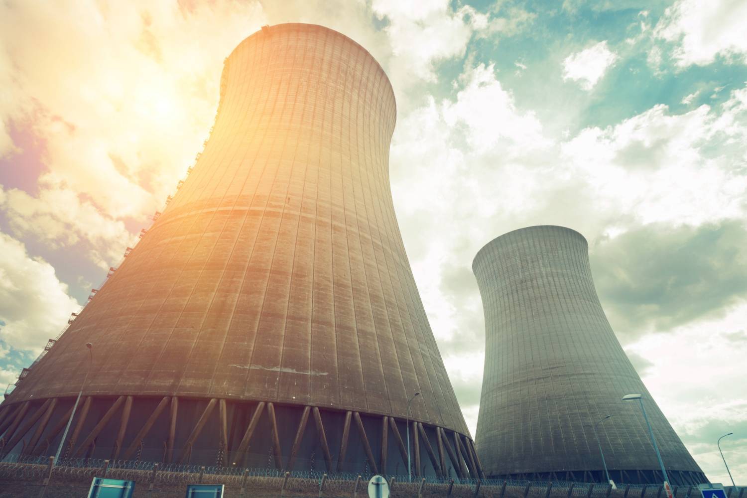 Two large cooling towers against a cloudy sky with sunlight filtering through, surrounded by fences and lampposts.