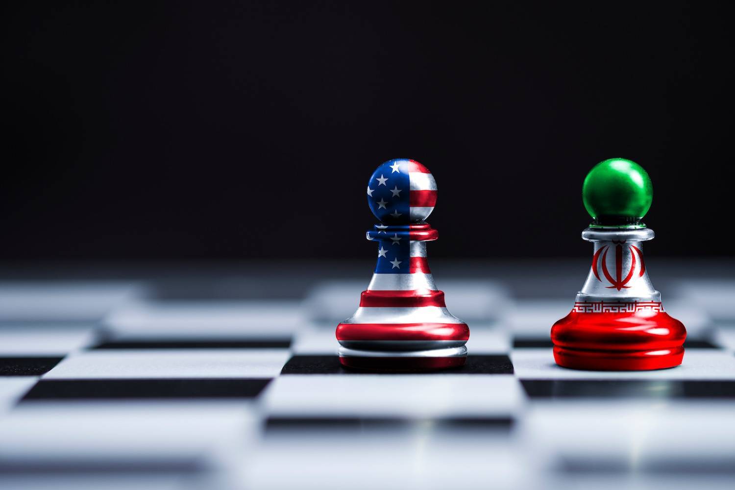 Two pawns with American and Iranian flags stand on a chessboard against a dark background.