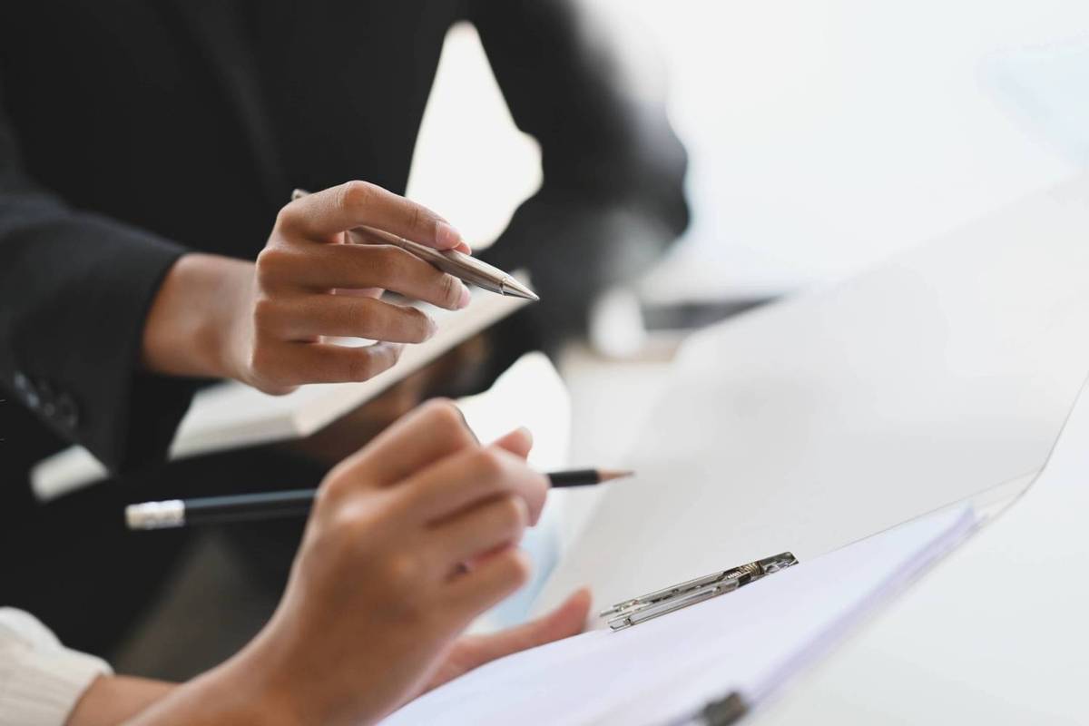 Two people in suits hold pens over a clipboard with papers in a bright office setting.
