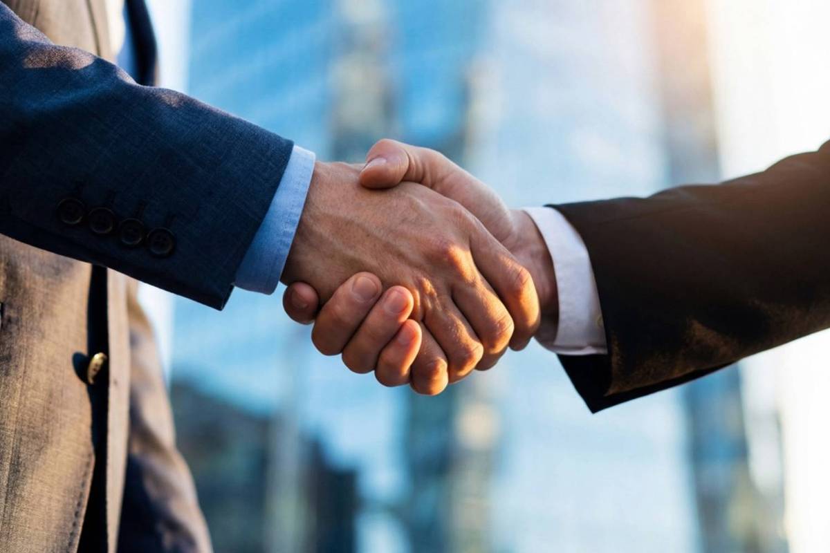 Two people in suits shaking hands in front of a tall, glass-covered building.