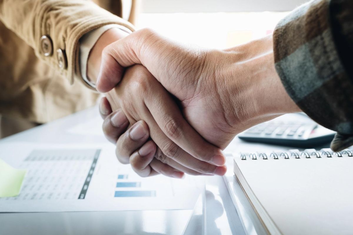 Two people shaking hands over a desk with documents and a notebook.