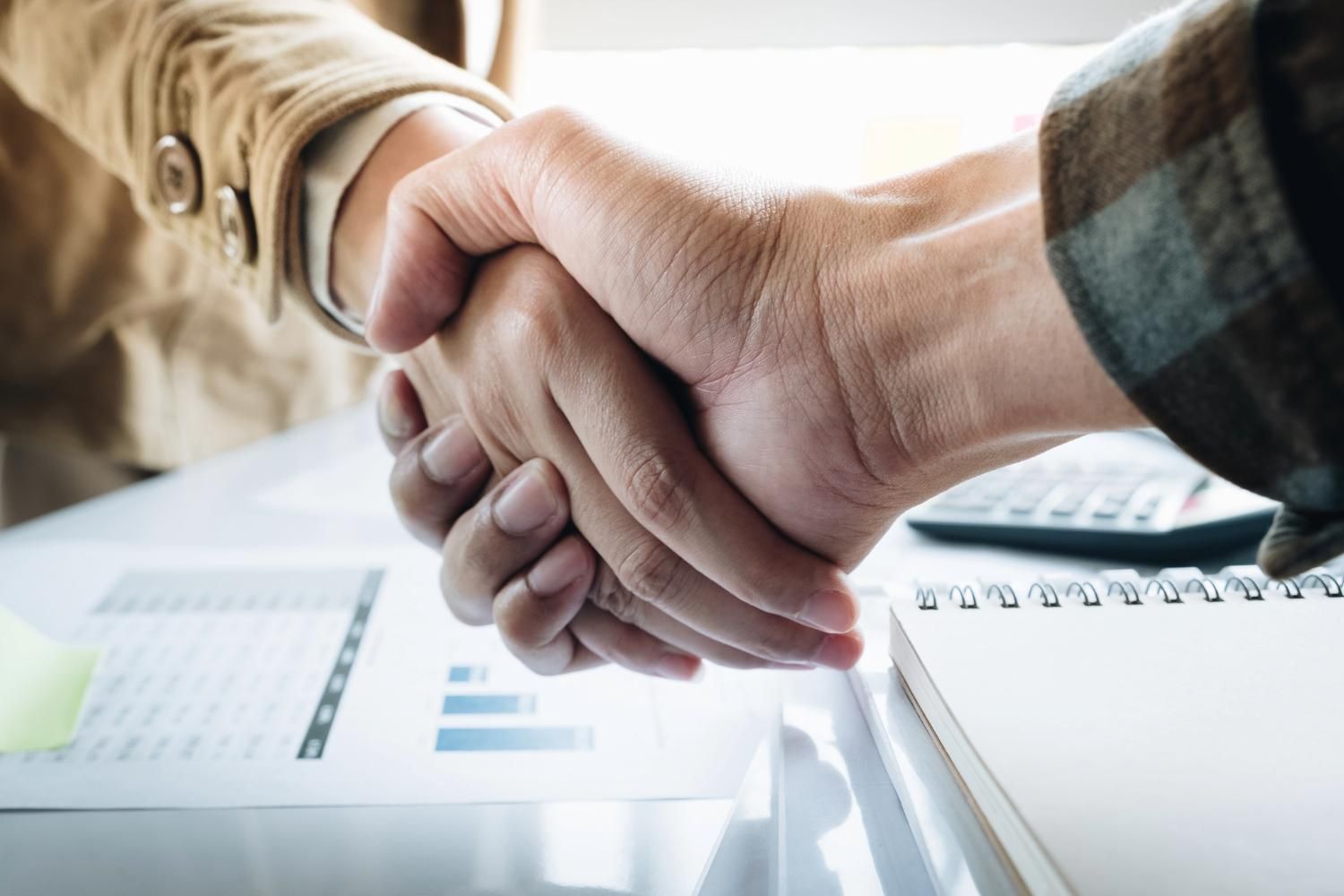 Two people shaking hands over a desk with documents and a notebook.
