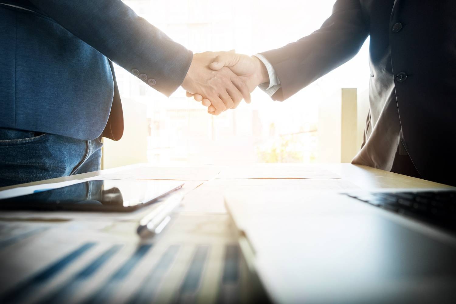Two people shaking hands over a desk with papers and a tablet.