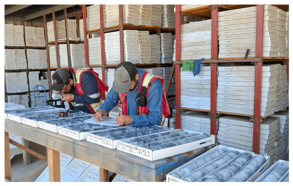 Two Silver Dollar Resources workers examine rock samples on a wooden table, with shelves full of boxes behind them.