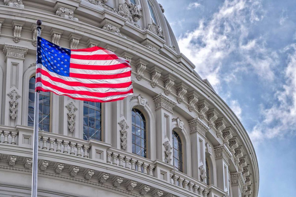 U.S. flag waving near the ornate Capitol building dome against a partly cloudy sky.