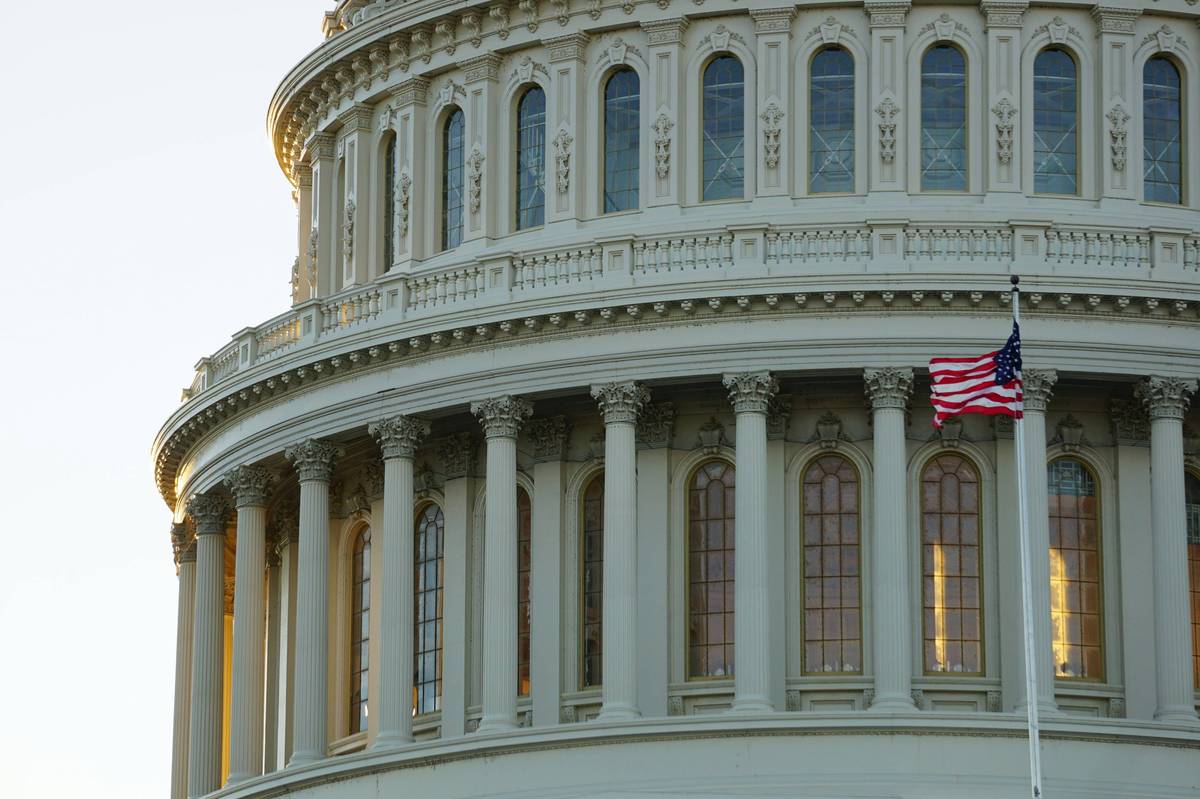 US Capitol dome with intricate columns and a waving American flag in the foreground.